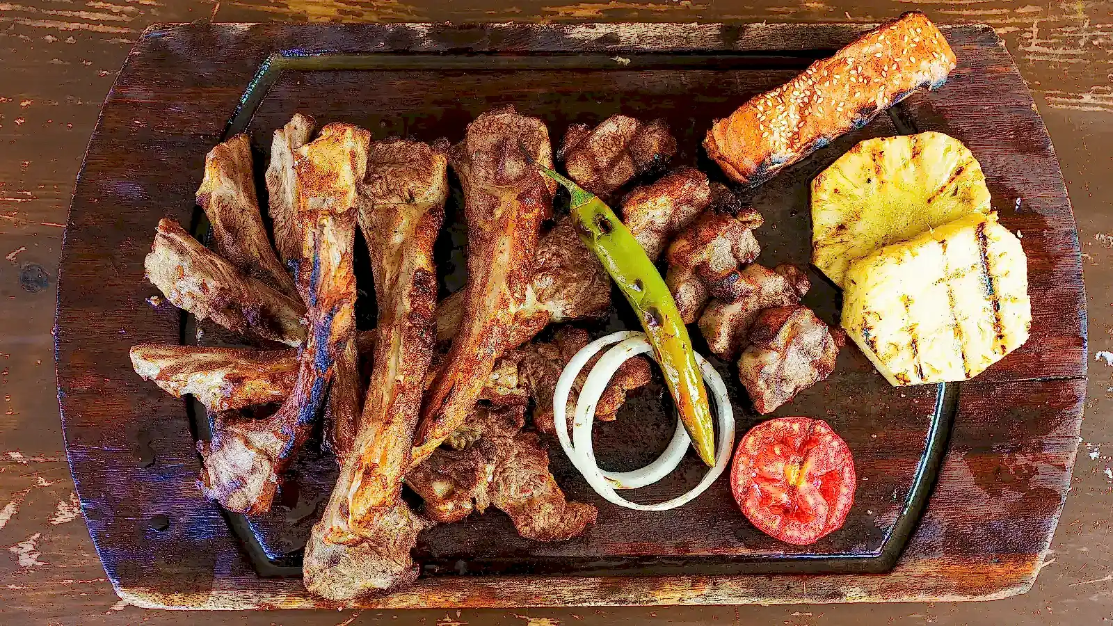 Kömür ateşinde pişirilmiş kuzu kafes ızgara, yanında közlenmiş biber, domates, halka soğan ve ananas dilimiyle döküm tabakta sunum – Adana Dede Kebap Steak Burger Tatlı Kasap